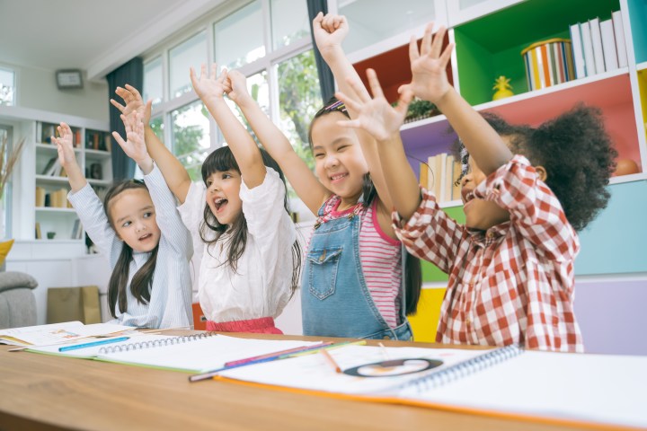 Four children sitting at a table, raising arms and smiling in a colorful classroom.