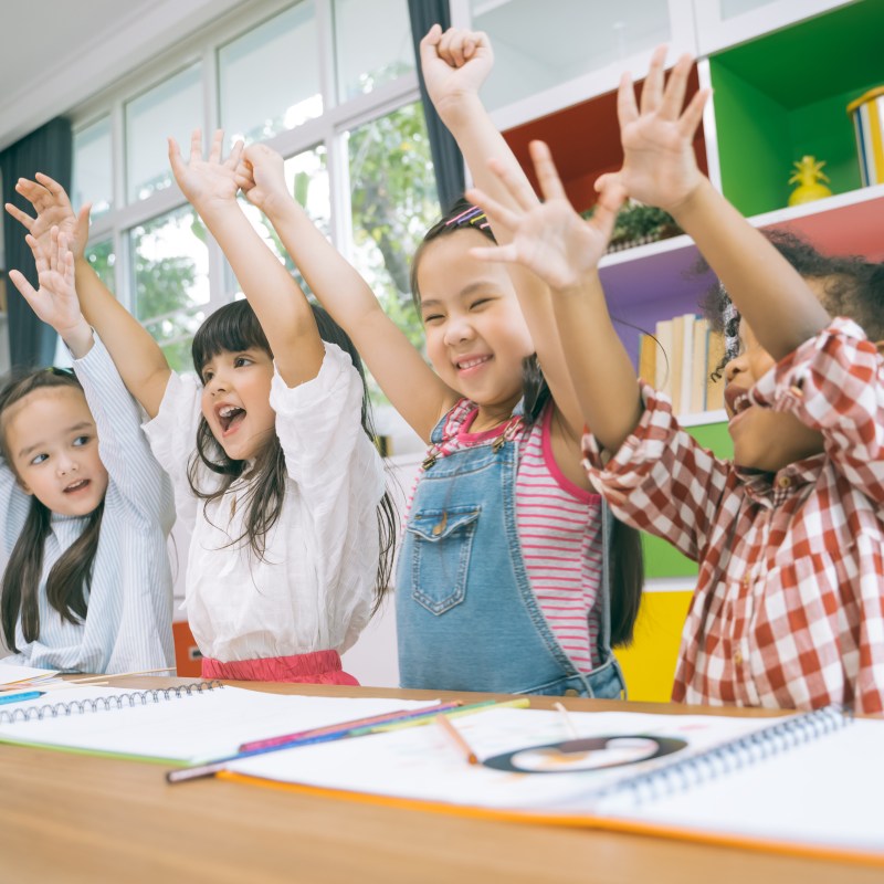 Four children sitting at a table, raising arms and smiling in a colorful classroom.