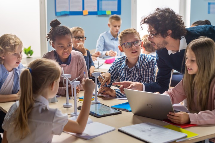 Teacher with students discussing a project using wind turbine models and laptops in a classroom.