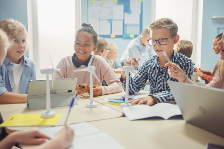Children in a classroom working on a wind turbine project at a table with laptops and tablets.