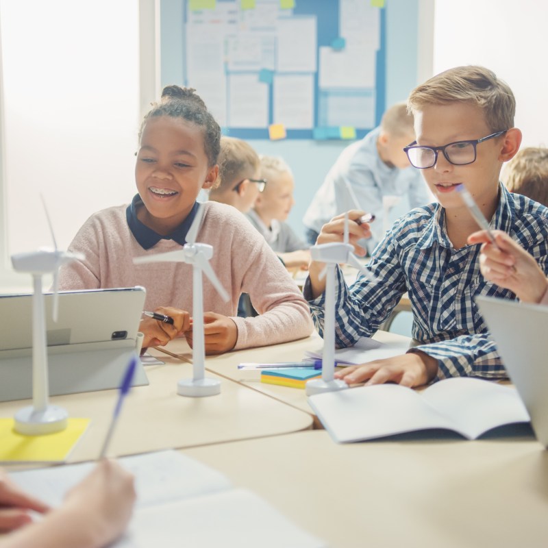Children in a classroom working on a wind turbine project at a table with laptops and tablets.
