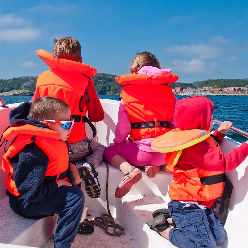 Four children in life jackets on a boat, looking towards the sea and distant shore.