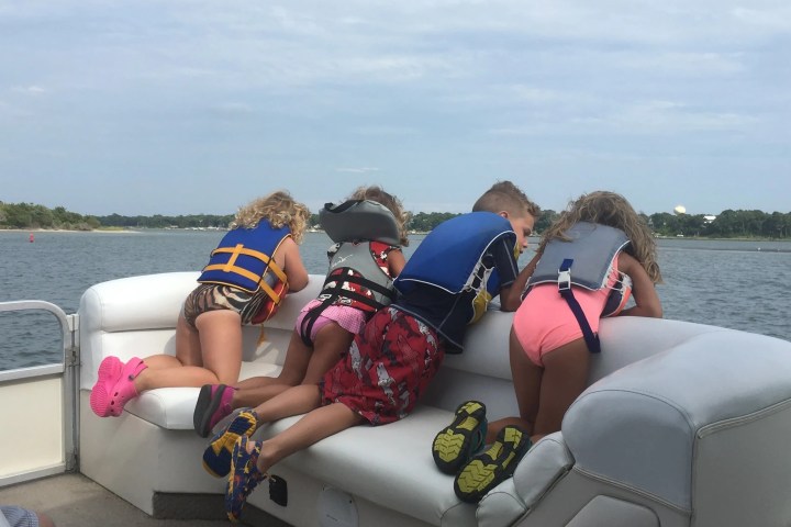 Four children in life jackets leaning over a boat railing, watching the water.