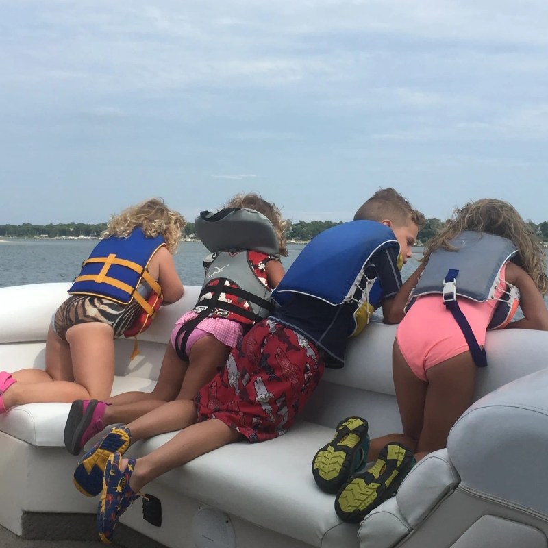Four children in life jackets leaning over a boat railing, watching the water.